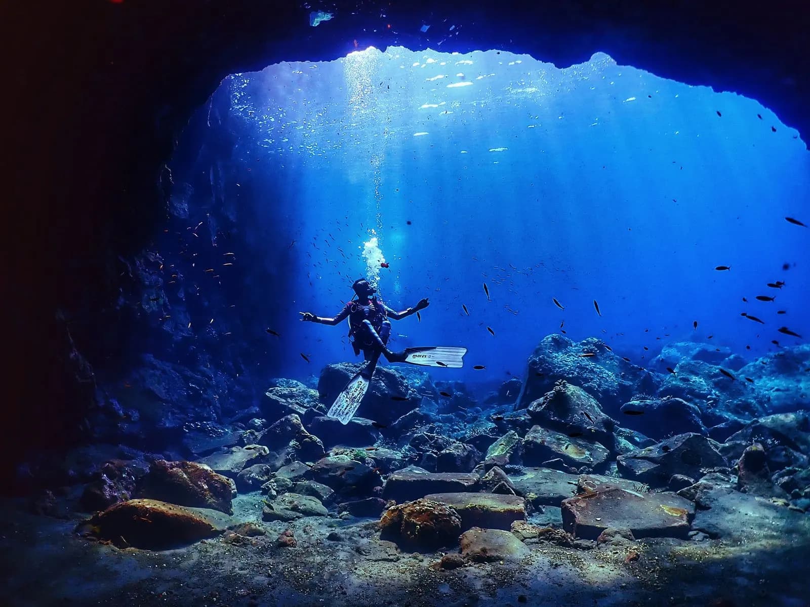 Diver in a sunlit underwater cavern with god rays and schooling fish